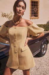 A woman wearing a light yellow, off-the-shoulder dress with large puffed sleeves. The dress features a row of oversized buttons down the front and a small flap pocket on the right side. She accessorizes with a chunky gold necklace and matching earrings.